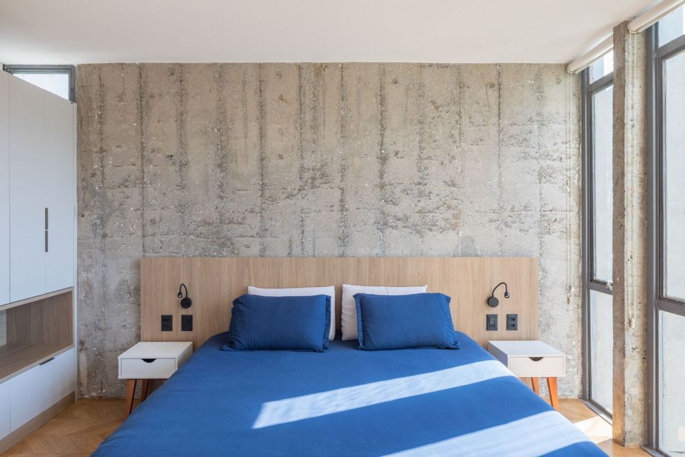 La chambre a un style similaire, avec un mur en béton apparent et le même motif de sol en chevron