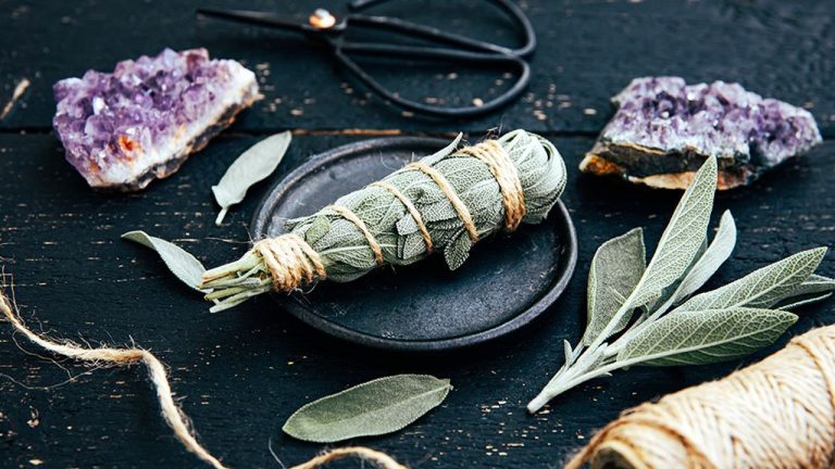 White homemade sage Salvia apiana smudge stick at home with homegrown sage leaves. Cotton string, vintage scissors and amethyst clusters for decoration on black wood background. Side view.